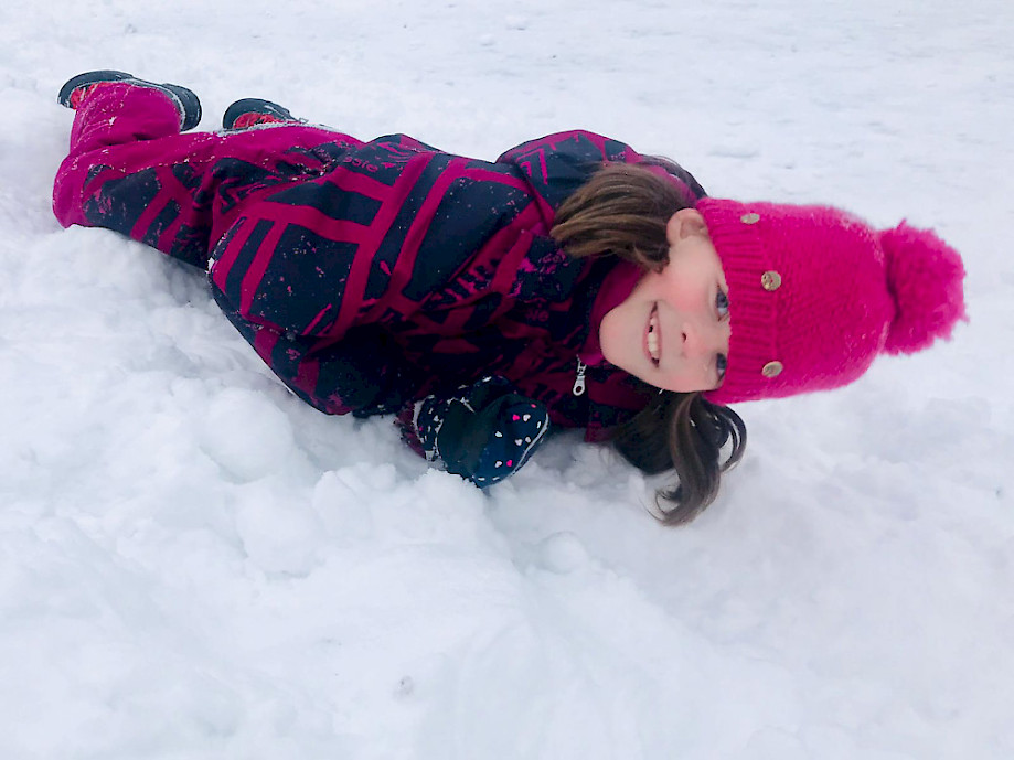 Pinkkipipoinen lapsi makaa toppavaatteissa lumessa ja virnistää kameralle.